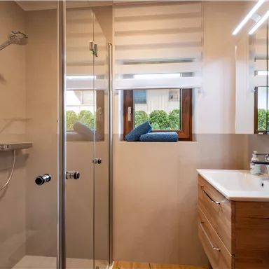 A modern bathroom with a glass shower and a wooden washbasin. The window offers a view of a green outdoor area.