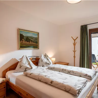 A cozy bedroom with two beds, colorful pillows, and a painting on the wall.  
Large windows overlooking the garden provide natural light.