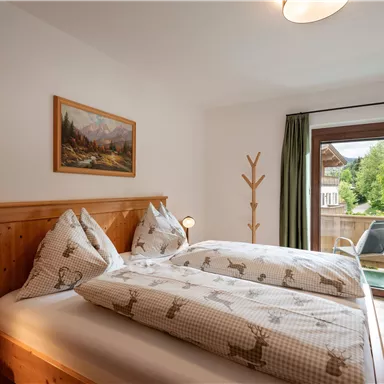 A cozy bedroom with a double bed and wooden details. Large windows provide a view of a green outdoor area.