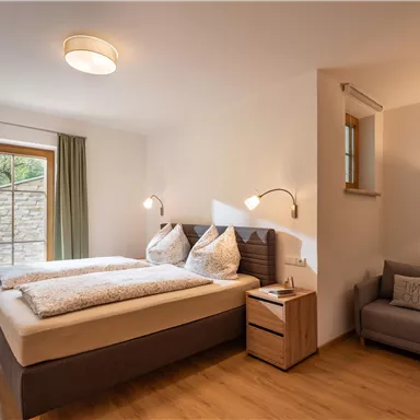 A modern bedroom with a double bed and two pillows. There is a cozy armchair and large windows with curtains.