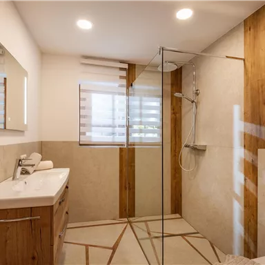 A modern bathroom with a glass shower and wooden details. The walls are designed to be light and welcoming, with a large mirror above the sink.