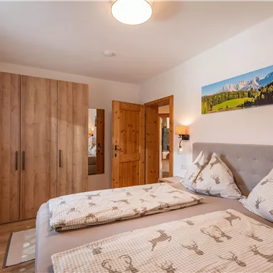 A cozy bedroom with a double bed and a wooden dresser. The walls are light, and a large window provides plenty of daylight.