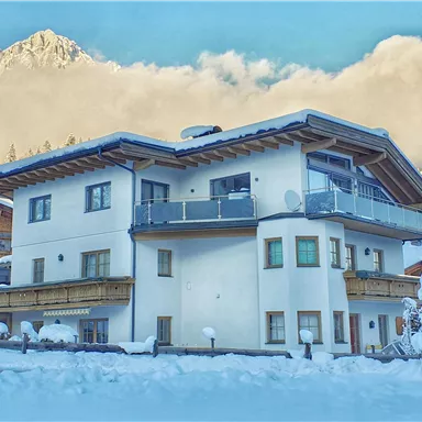 A modern house in winter, surrounded by snow. In the background, mountains and a blue sky can be seen.