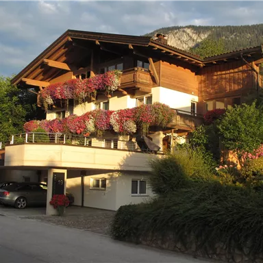A charming wooden house with colorful flowers on the balcony. In the foreground, cars and plenty of greenery can be seen.