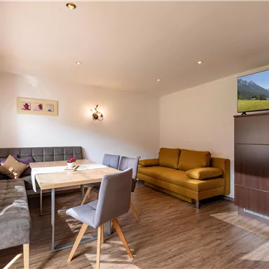 Ein helles Wohnzimmer mit einem Esstisch, einer gemütlichen Couch und einem Fernseher. Die Räume sind modern und freundlich eingerichtet.