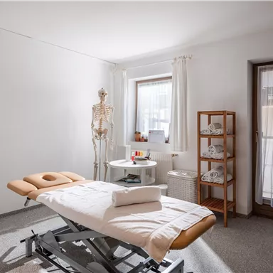 A bright room for physiotherapy treatments with a treatment table, a skeleton, and a shelf for towels. The large windows provide plenty of daylight.