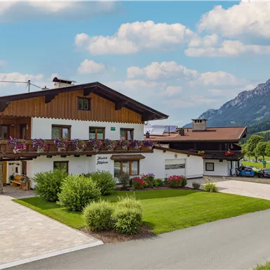 A charming country house with flower balconies, surrounded by green meadows and mountains. The sky is blue with a few clouds.