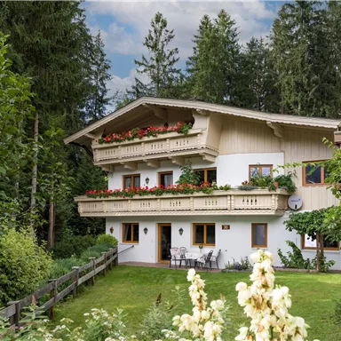 Ein charmantes Holzhaus mit Balkon, umgeben von grünen Bäumen und bunten Blumen. Die Atmosphäre ist ruhig und einladend.