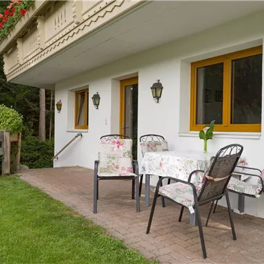 Ein gemütlicher Sitzbereich mit einem Tisch und Stühlen ist vor einem Wohnhaus im Grünen angeordnet. Der Rasen ist gepflegt und es gibt einige Pflanzen in der Nähe.