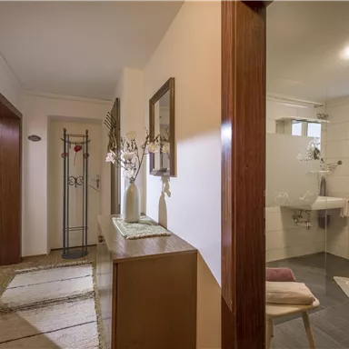 A bright and welcoming hallway with a wooden cabinet and modern mirrors. The walls are painted in a neutral shade, and there is a small washing area.