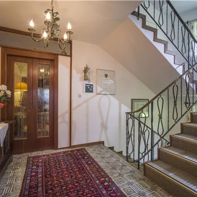 An inviting hallway with a carpet and a beautiful staircase. The walls are designed in warm colors, and the light comes from a decorative chandelier.