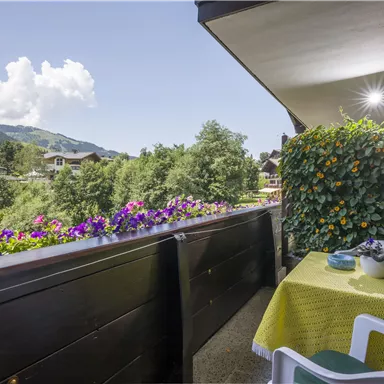 A cozy balcony with a beautiful view of mountains and trees. A table with a yellow tablecloth and a flower box creates an inviting atmosphere.
