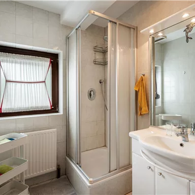 Ein modernes Badezimmer mit einer Dusche und einem Waschbecken. Es gibt eine helle Wandgestaltung und einen großen Fenster mit Rollo.