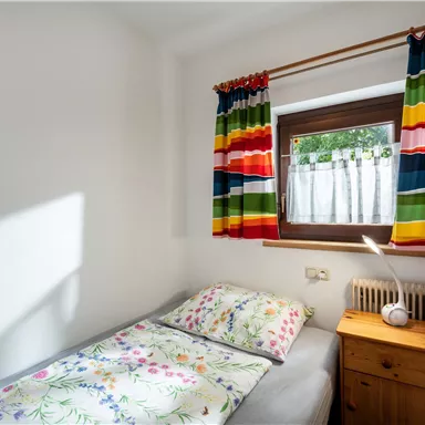 Ein kleines, helles Zimmer mit einem Einzelbett und bunten Vorhängen. Auf dem Nachttisch steht eine Lampe und das Fenster lässt viel Licht herein.