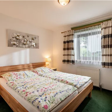 Ein gemütliches Schlafzimmer mit einem Doppelbett und bunten Bettdecken. Das Zimmer hat große Fenster, die Tageslicht hereinlassen, und eine moderne Dekoration.