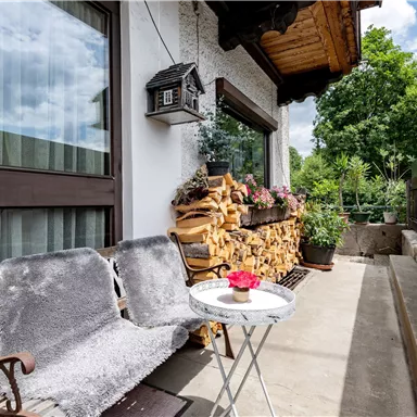 Ein gemütlicher Balkon mit Holzstapel, einer kleinen Tisch und einer Bank. Grünpflanzen und eine schöne Aussicht auf die Natur ergänzen die entspannte Atmosphäre.