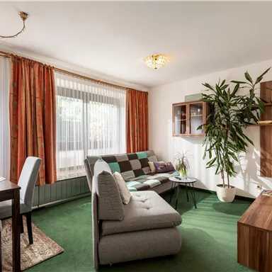 Ein helles Wohnzimmer mit einem gemütlichen Sofa und einem Essbereich. Große Fenster sorgen für viel Tageslicht und eine grüne Pflanze bringt Frische in den Raum.