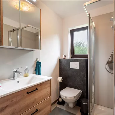 Ein modernes Badezimmer mit einer Dusche, einem weißen Waschtisch und Holzverkleidung. Die Wände sind hell und es gibt ein Fenster, das Tageslicht hineinlässt.