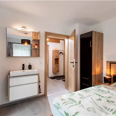 Ein modernes Schlafzimmer mit einem komfortablen Bett und einem großen Spiegel. Im Hintergrund ist ein Badezimmerbereich sichtbar, der eine Sauna enthält.