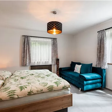 Ein modernes Schlafzimmer mit einem Doppelbett und einer gemütlichen Couch. Helle Vorhänge und eine stilvolle Lampe sorgen für eine einladende Atmosphäre.