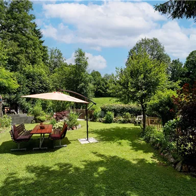 Ein schöner Garten mit grüner Wiese und vielen Bäumen. Im Vordergrund stehen ein Tisch und Stühle unter einem Sonnenschirm.