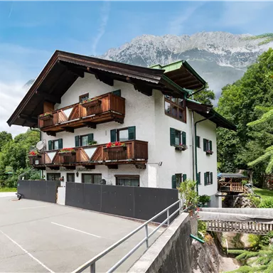 Ein hübsches, mehrstöckiges Haus mit Balkon und grüner Umgebung. Im Hintergrund sind Berge und ein blauer Himmel zu sehen.