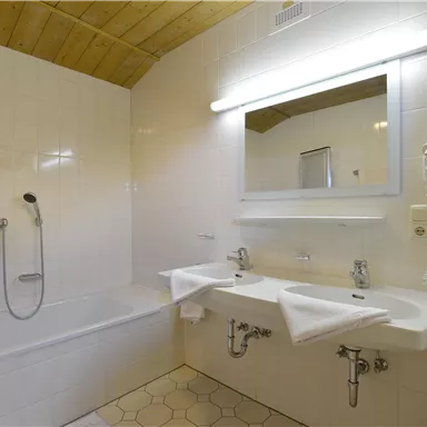 Ein modernes Badezimmer mit einer Badewanne und zwei Waschbecken. Es hat helle Fliesen und eine große Spiegelleuchte.