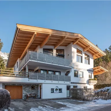 A modern, multi-story house with a large balcony and a wooden clad roof. It is located in a snow-covered environment with green trees in the background.