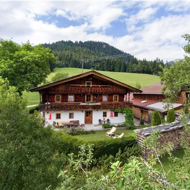 A traditional farmhouse surrounded by green meadows and trees. In the background, gentle hills and a blue sky can be seen.