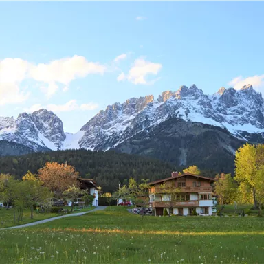 Eine malerische Landschaft mit majestätischen Bergen und schneebedeckten Gipfeln. Im Vordergrund sind grüne Wiesen und traditionelle Häuser zu sehen.