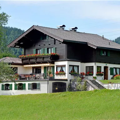 Ein traditionelles Haus im alpenländischen Stil, umgeben von einem grünen Garten. Im Hintergrund sind sanfte Hügel und Bäume sichtbar.
