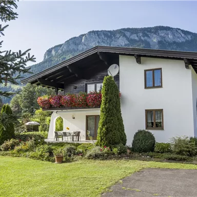 Ein schönes Haus umgeben von einem gepflegten Garten und grünen Bäumen. Im Hintergrund sind majestätische Berge zu sehen.
