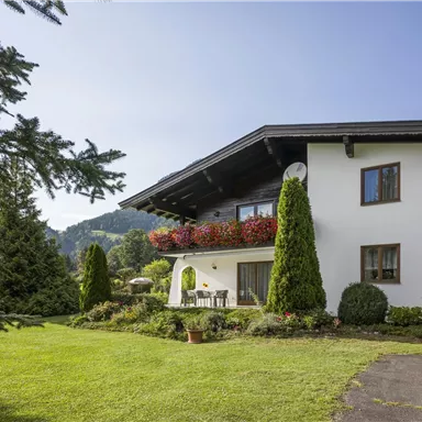 Ein hübsches Haus mit einem gepflegten Garten und Bäumen. Im Hintergrund sind sanfte Hügel zu sehen.