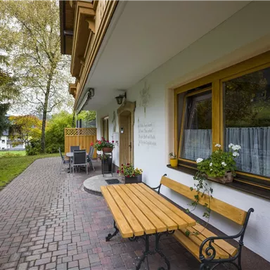 Ein gemütlicher Außenbereich mit einem Holzbank und Blumen vor einem Haus. Umgeben von Grünflächen und Bäumen, strahlt die Szene Ruhe und Wärme aus.