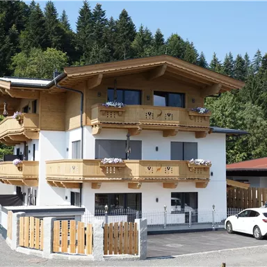 Ein modernes Gebäude im alpinen Stil mit Balkonen aus Holz. Umgeben von grünen Bäumen und einem Parkplatz.
