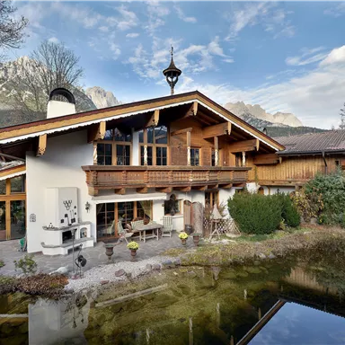 A beautiful wooden house in Alpine style with large windows and a well-kept garden. In the foreground, a small pond can be seen.