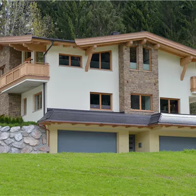 A modern house with a wooden and stone facade in a green environment. The large meadow and the trees in the background give the image a peaceful atmosphere.