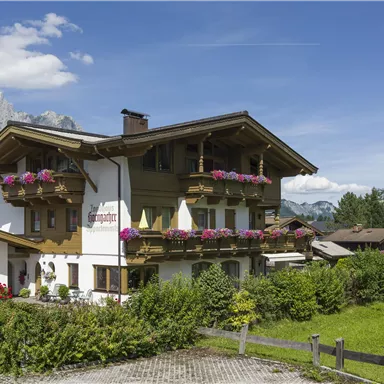 A traditional alpine house with balconies full of flowers, surrounded by green landscape. In the background, majestic mountains and a blue sky can be seen.