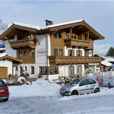 A rustic building in alpine style, surrounded by snow. In front of the house are two cars and in the background, mountains are visible.
