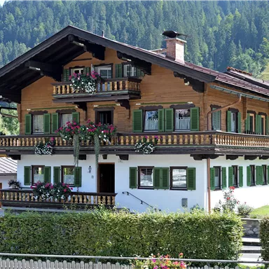 A traditional, multi-story wooden house with green shutters and floral decorations. Surrounded by a well-maintained hedge and the picturesque Alpine landscape.