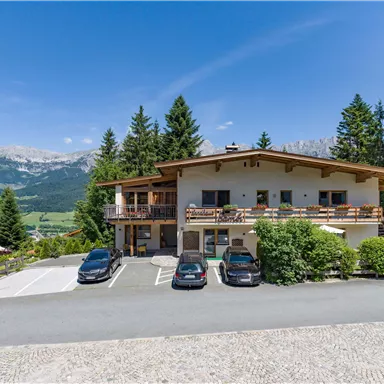 Ein Gebäude im alpinen Stil, umgeben von Bäumen und Bergen. Vor dem Haus stehen mehrere Autos auf einem gepflasterten Parkplatz.