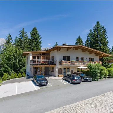 Ein charmantes Gebäude in den Bergen mit umgebenden Bäumen. Die Straße vor dem Haus ist mit mehreren parkenden Autos gesäumt.