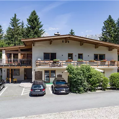 Ein modernes Haus inmitten von Bäumen mit einem klaren blauen Himmel. Vor dem Gebäude stehen mehrere Autos auf einem befestigten Parkplatz.