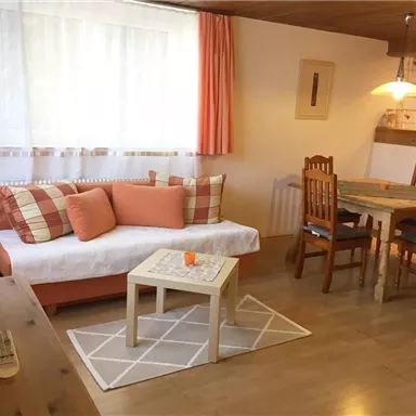 Ein gemütlicher Wohnraum mit einer hellen Couch und einem Holz-Esszimmertisch. Große Fenster sorgen für viel Licht und eine einladende Atmosphäre.