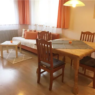 Ein helles Wohnzimmer mit einem gemütlichen Sofa und einem Tisch mit Stühlen. Große Fenster lassen viel Licht herein, und die orangefarbene Dekoration schafft eine einladende Atmosphäre.