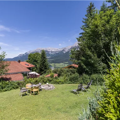 Eine idyllische Landschaft mit einem grünen Garten und Blick auf Berge. Im Vordergrund stehen Liegestühle und eine Feuerstelle.