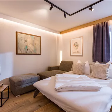 Ein modernes Zimmer mit einem bequemen Bett und einer gemütlichen Sitzecke. An der Wand hängen Bilder und der Raum hat große Fenster mit Vorhängen.