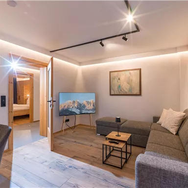 Ein modernes Wohnzimmer mit einer grauen Couch und einem Fernseher. Der Raum ist hell und freundlich gestaltet, mit warmem Licht und Holzdetails.