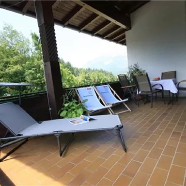 Ein gemütlicher Balkon mit Liegen, Stühlen und einem Tisch. Im Hintergrund sind grüne Bäume und die Berge zu sehen.