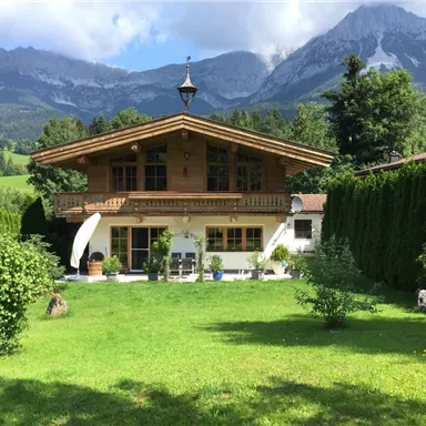 Ein schönes, traditionelles Holzhaus umgeben von grünem Rasen und Bäumen. Im Hintergrund erheben sich majestätische Berge unter einem klaren Himmel.